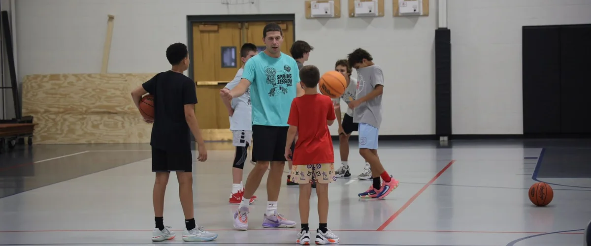A male coach in a teal shirt instructing young boys with basketballs on an indoor court. A male coach in a teal shirt instructing young boys with basketballs on an indoor court.