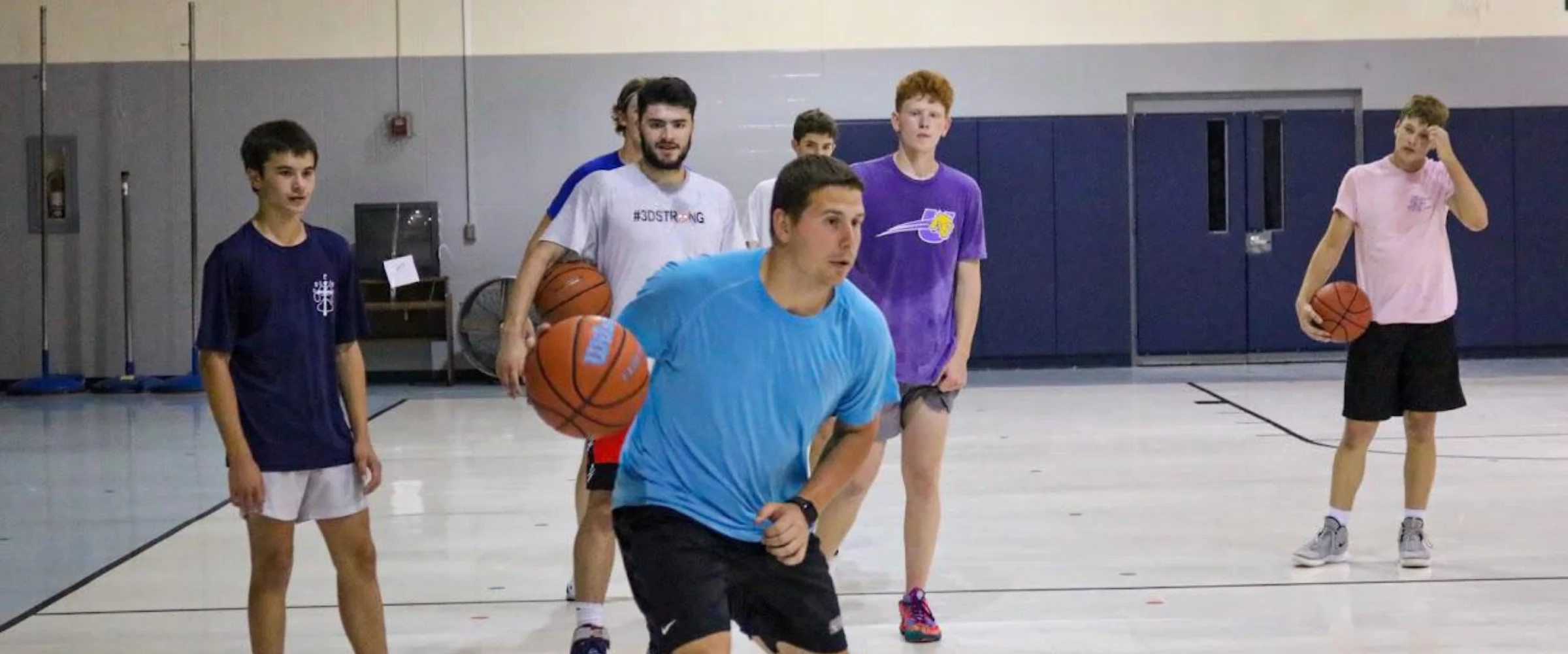 A group of young men training with basketballs in a gymnasium, led by a man dribbling in the foreground. A group of young men training with basketballs in a gymnasium, led by a man dribbling in the foreground.