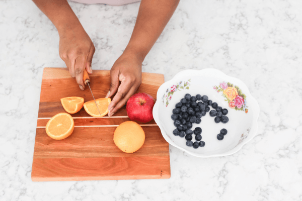 A person slices an orange on a wooden cutting board beside a red apple and a bowl of blueberries, illustrating simple, practical meal preparation for someone living with MS.