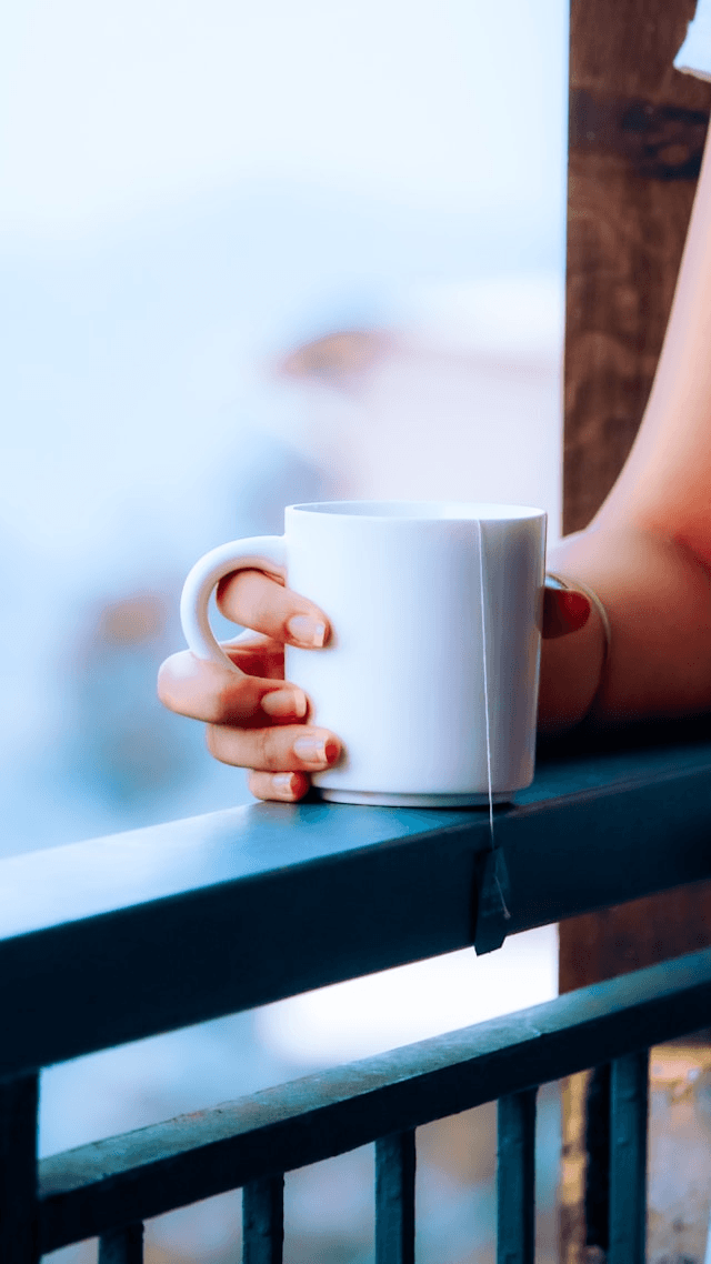 A hand holding a white mug of tea on a balcony railing with a blurred background. A hand holding a white mug of tea on a balcony railing with a blurred background.