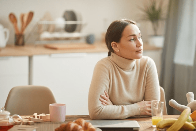 A person wearing a beige turtleneck sweater sits at a table with their arms crossed, looking to the side. 