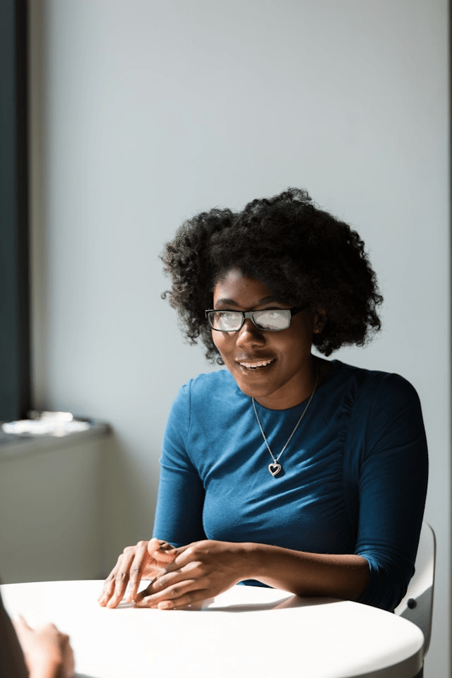 Woman with curly hair and glasses sits at a table, smiling and talking with someone seated across from her.