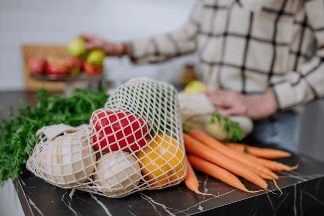 A reusable mesh bag filled with produce including red bell peppers, onions, and oranges, next to carrots and leafy greens on a countertop. A reusable mesh bag filled with produce including red bell peppers, onions, and oranges, next to carrots and leafy greens on a countertop.