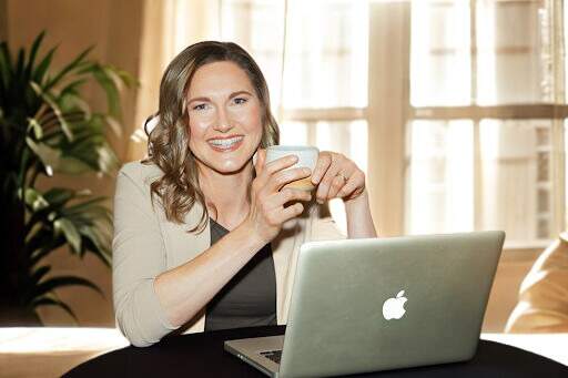 Linda Bartholme smiling behind her laptop by a bright window, designing corporate mental fitness programs.