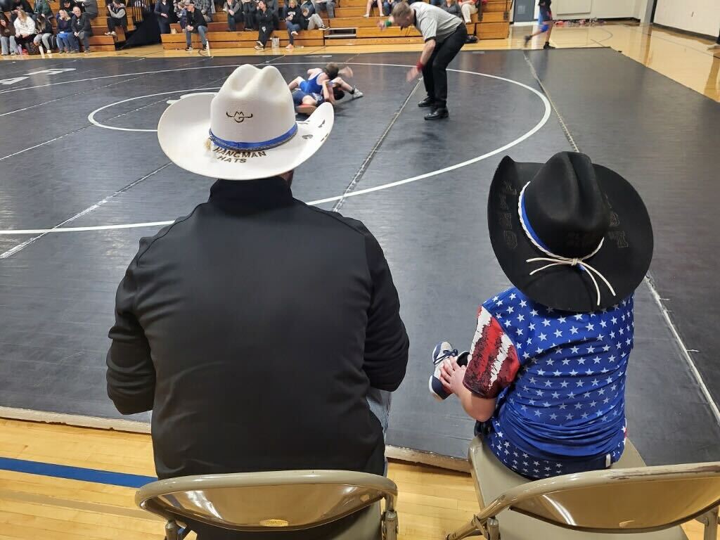 The owner of Hangman Hats coaching wrestling in his cowboy hat, with a wrestler wearing the Grit Hat courtside