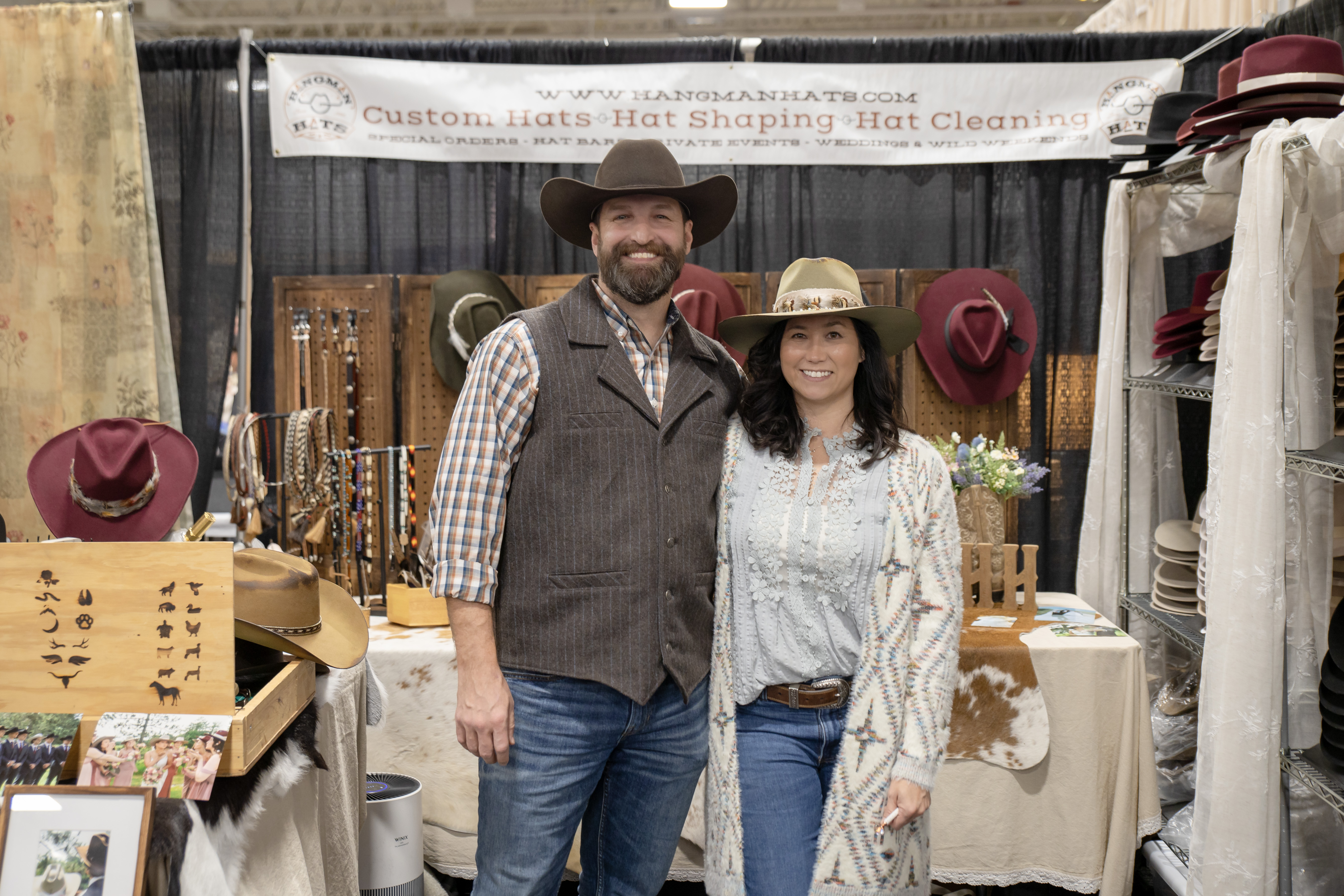 Jen and the owner of Hangman Hats standing in front of their booth at a wedding show, both wearing custom hats