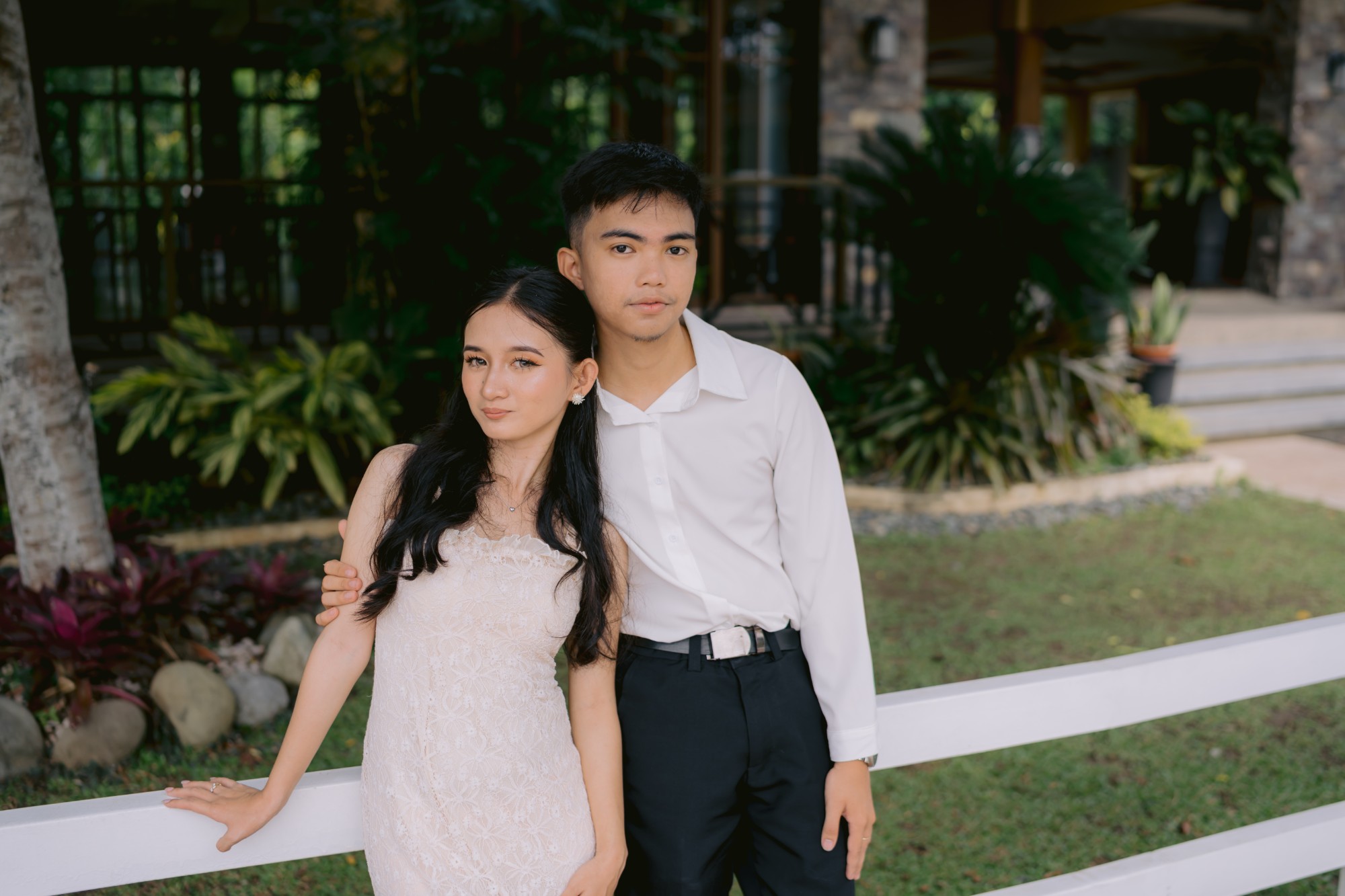The couple leaning together against a white garden fence, she in a blush lace dress and he in a white shirt and black trousers