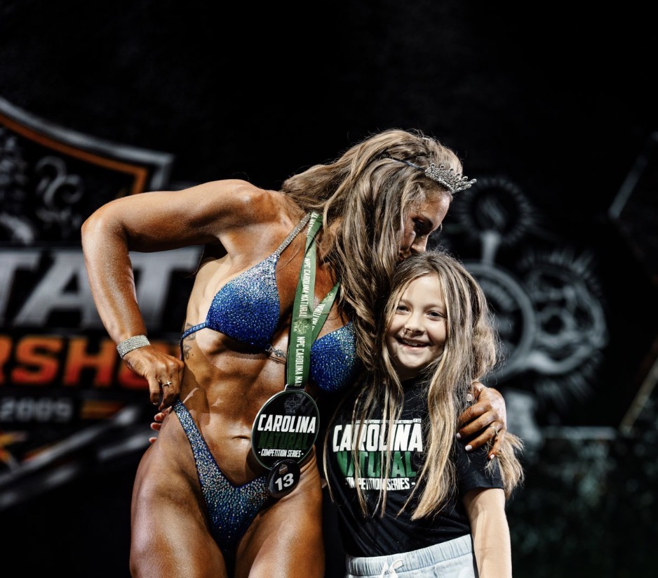 Cam Brown on stage at the NPC Upstate Natural with her daughter, wearing her Carolina Natural medal