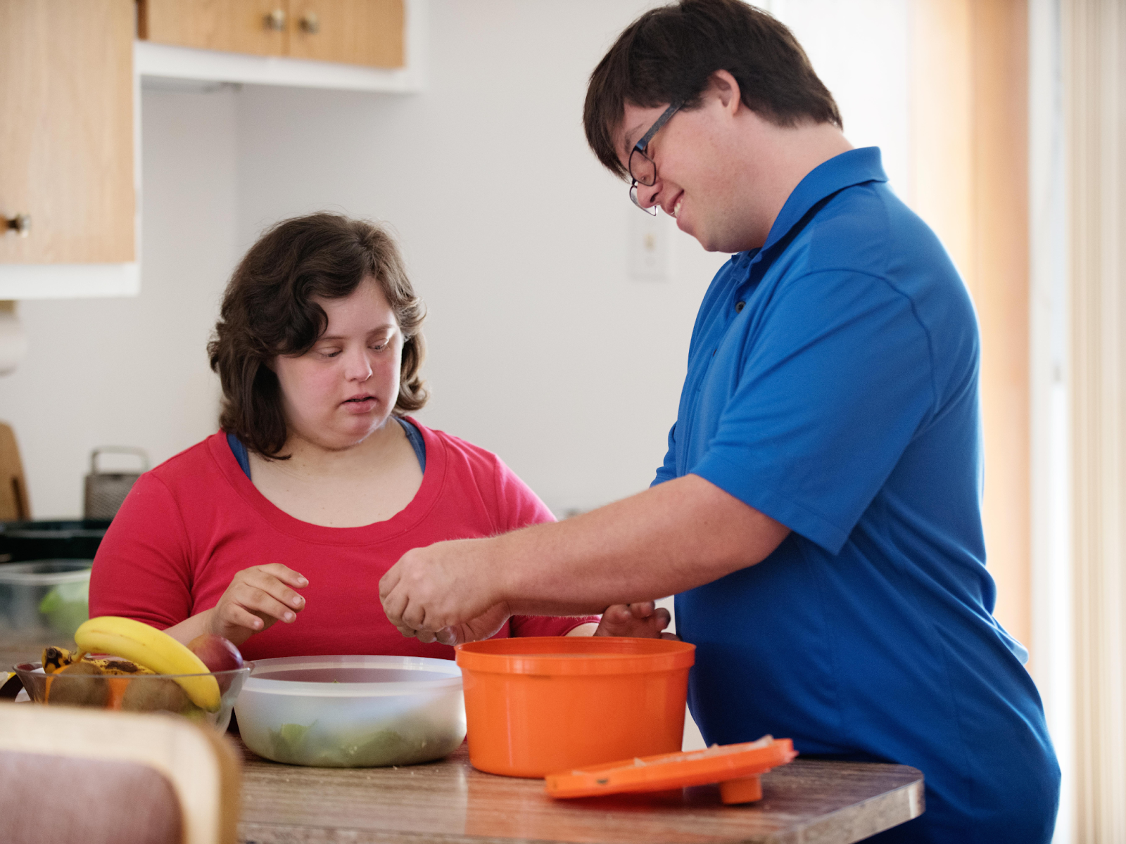 An adult preparing food in a kitchen with another person nearby.