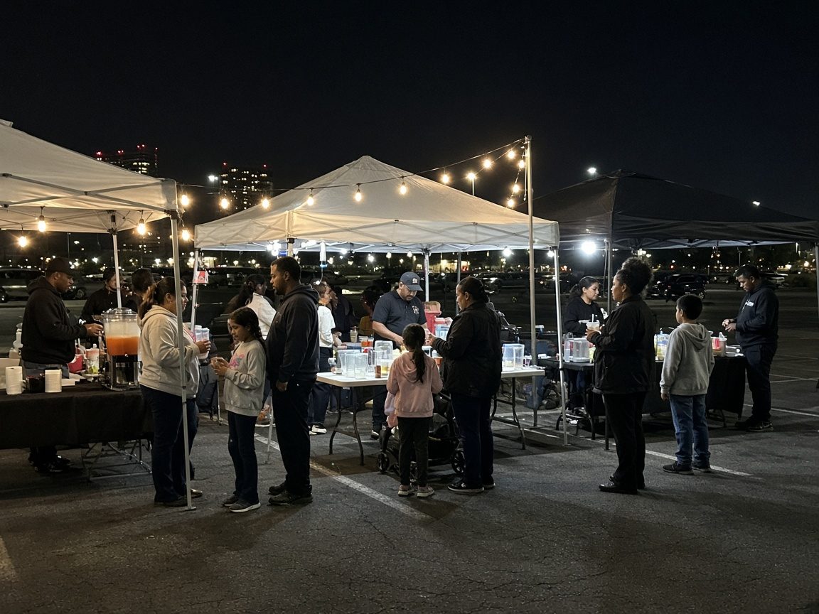 Families gathered at food vendor canopies with folding tables in a parking lot under string lights at the Dark Rooms event.