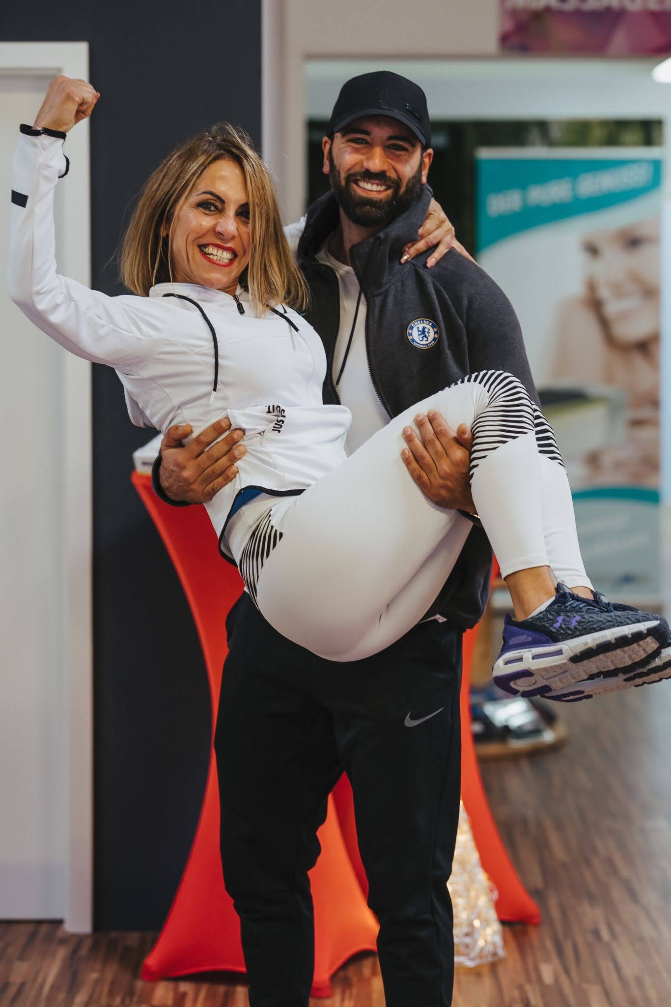 Trainer trägt lachend eine Kundin durchs Studio