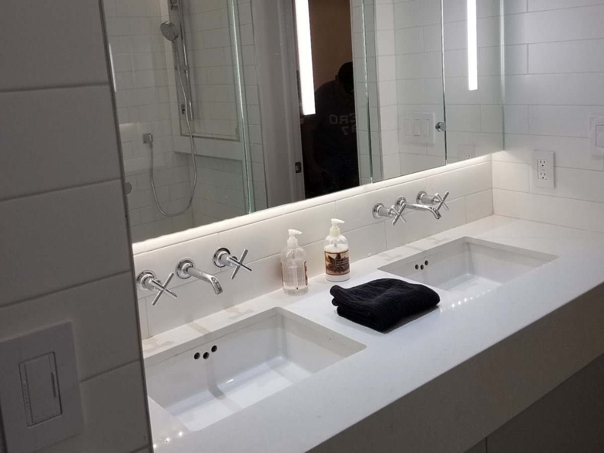 A contemporary bathroom sink with a double vanity setup, featuring wall-mounted faucets, a large mirror, and ample lighting, designed for a modern aesthetic.