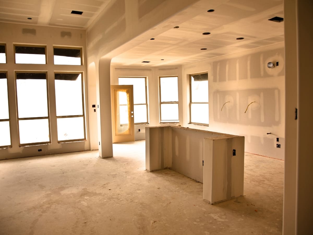 An open-concept living area with large windows and freshly installed sheetrock, showcasing a bright, spacious interior under construction.