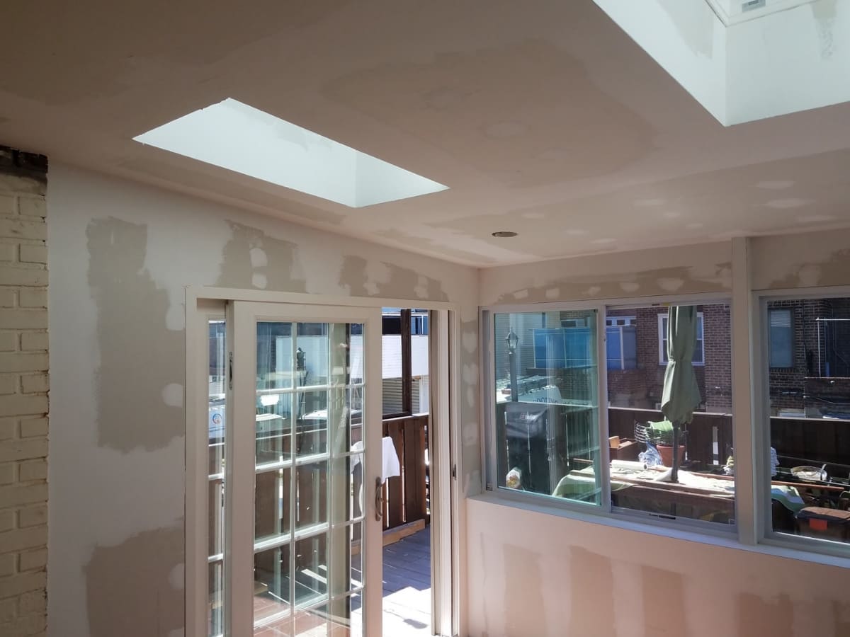 Sunroom under construction with drywall installation, featuring large windows and a skylight.