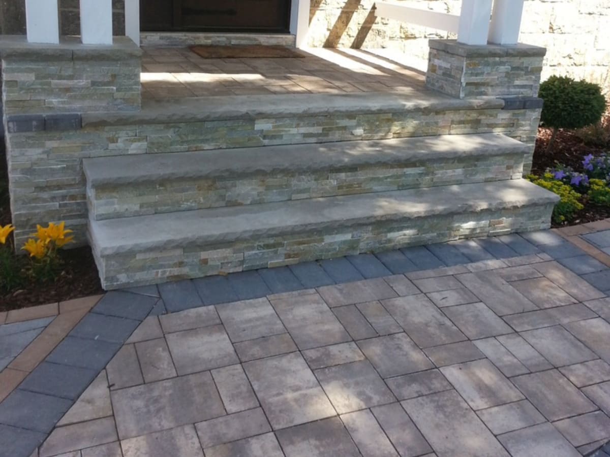 A beautifully designed entryway featuring stone paver steps leading up to the front porch, with matching stone accents on the columns and vibrant landscaping.