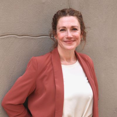 Kate Calhoun, co-founder of LLocalz and custom travel planner, standing in front of a gray wall with her arms behind her back, smiling warmly.