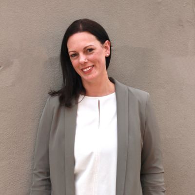 Denise Mackey, LLocalz co-founder and European travel specialist, smiling in front of a gray wall, wearing a green blazer and white shirt.