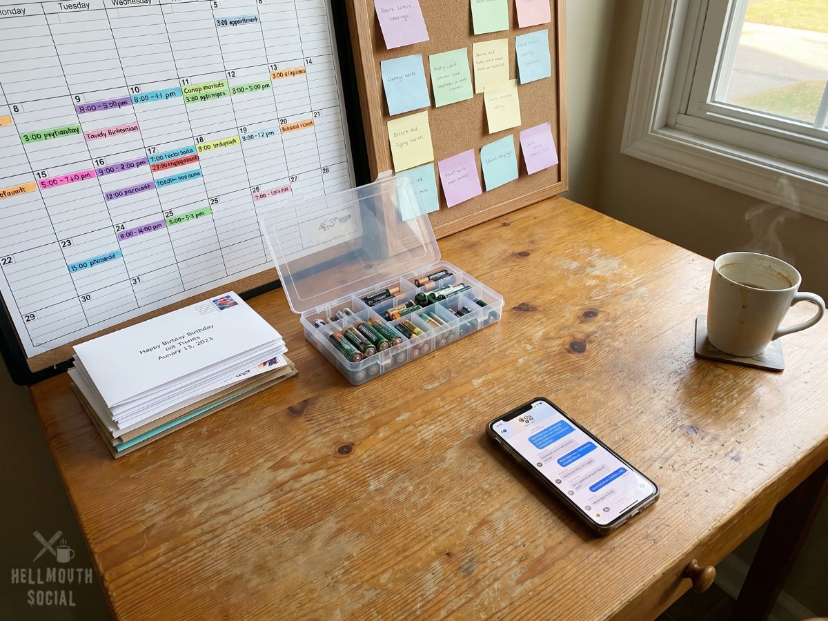 a kitchen table covered in color-coded calendars, sticky notes, birthday cards ready to be mailed, spare batteries, and a smartphone overflowing with group-chat notifications. Everything is perfectly organized except the rapidly cooling cup of coffee in the corner.