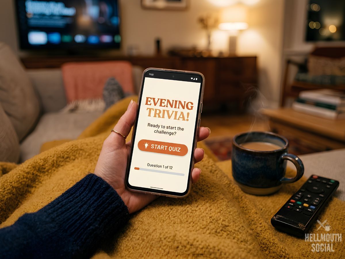 an inviting close-up of a hand holding a phone displaying a quiz start screen, coffee mug and TV remote nearby on a blanket. Warm evening living-room light, cozy and casual, shallow focus, welcoming editorial mood.