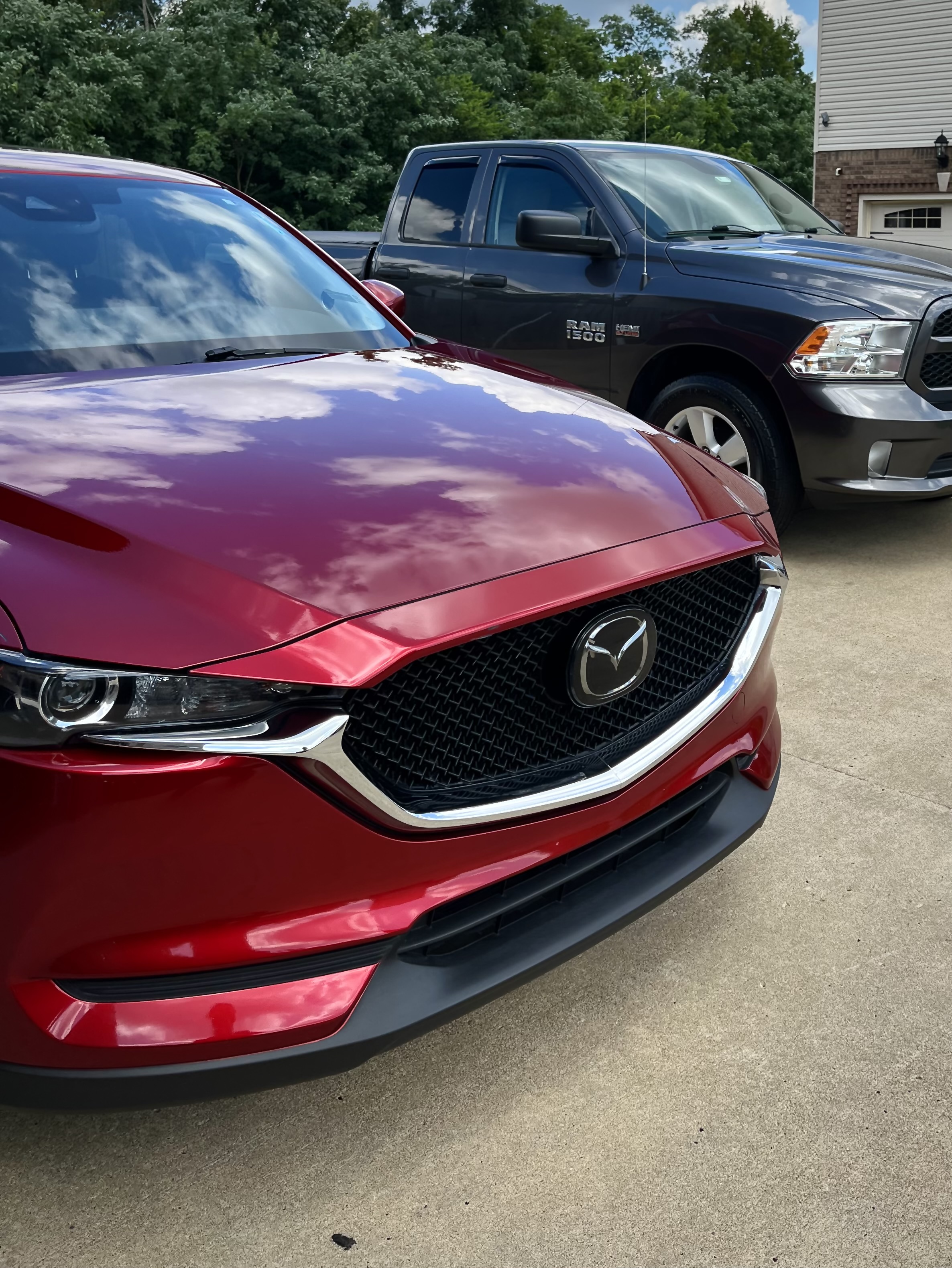 Close-up of the front end of a red Mazda CX-5 showing a glossy, freshly detailed hood and grille with a dark Ram 1500 truck parked behind it in a residential driveway.