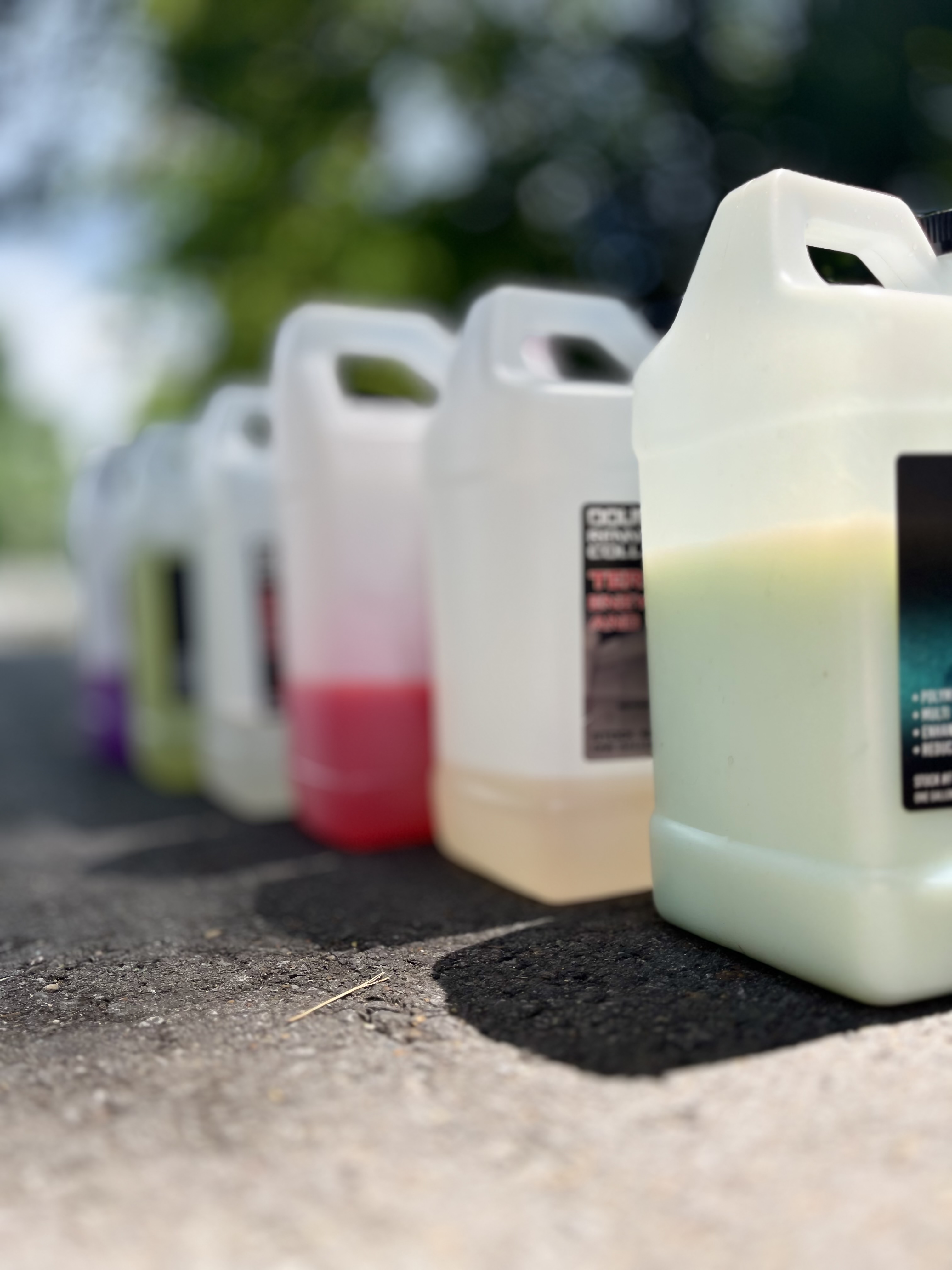 A row of colorful detailing chemical jugs lined up on pavement outdoors, representing the professional-grade products used by a mobile detailing service.