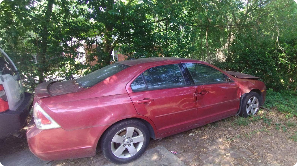 Red Ford Fusion sedan with front-end damage parked near wooded area and another vehicle.