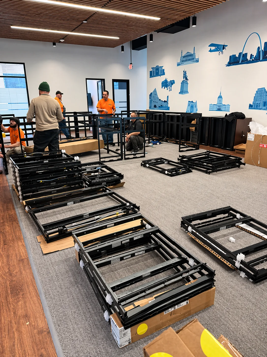 A team of five men in an office with a city mural on the wall, assembling black furniture frames.