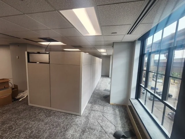 An empty office with light gray cubicles, gray patterned carpet, and large windows revealing a street view.