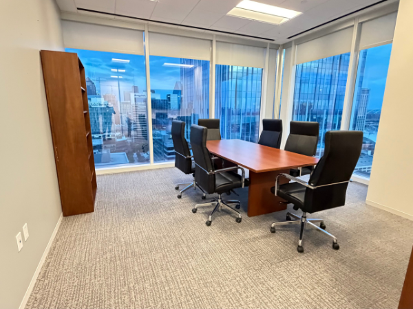 A modern office meeting room with a wooden table, black chairs, and floor-to-ceiling windows overlooking a city skyline.