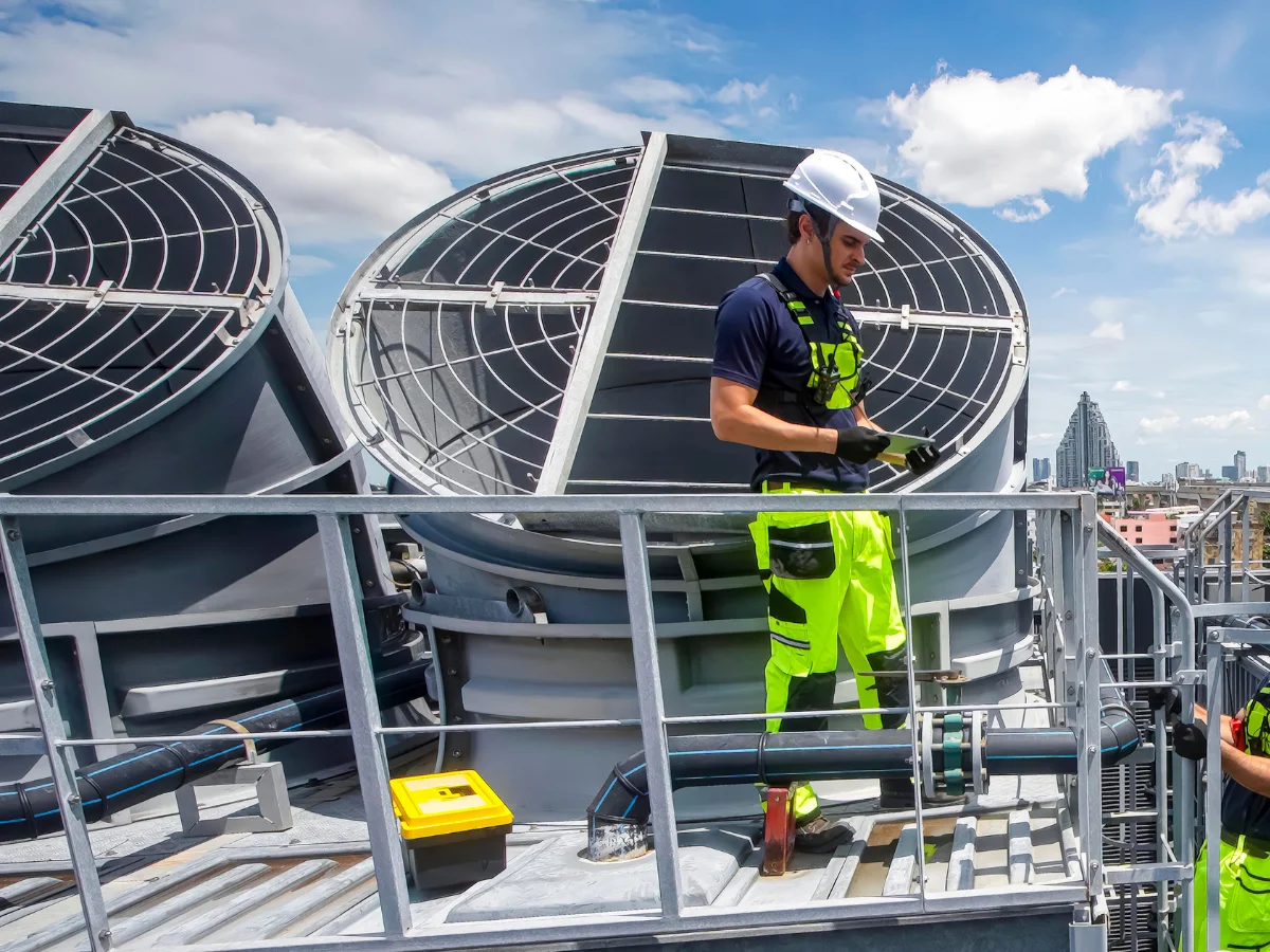 A man in safety gear inspecting industrial cooling towers on a rooftop with a city skyline in the background.