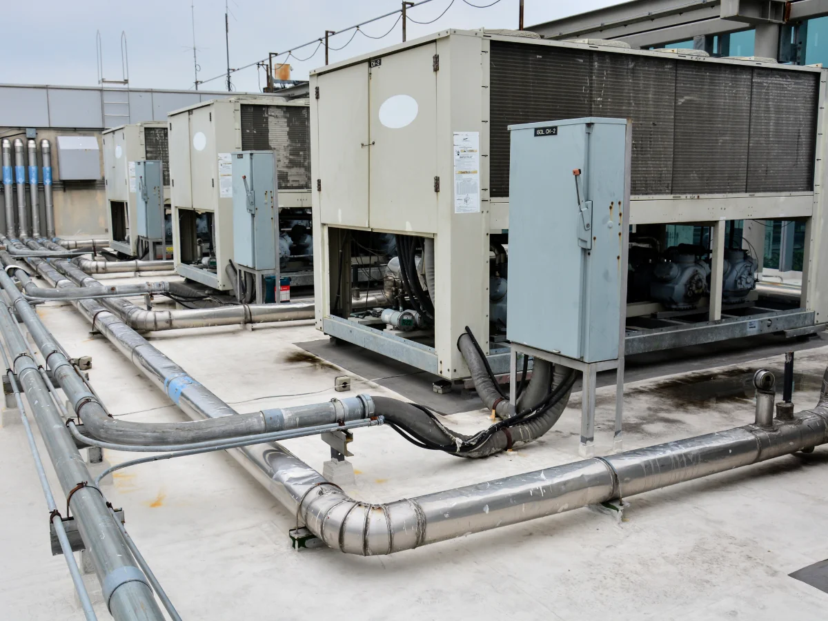 Multiple large industrial HVAC units and extensive silver piping on a commercial building's white rooftop under an overcast sky.