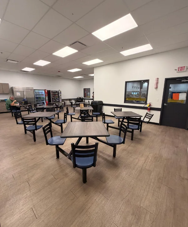 A wide interior view of a bright industrial cafeteria with numerous tables and chairs, a vending machine, and a man eating.