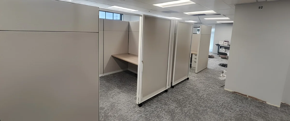 A wide shot of new office cubicle walls being installed on gray carpet, with unpacked boxes and a large window showing cars outside.