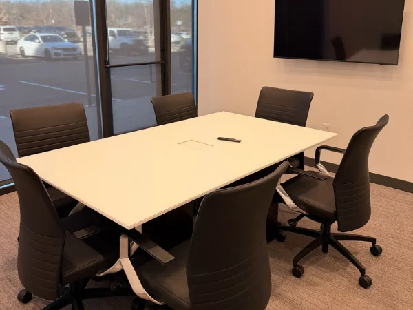 A modern conference room with a white table and black chairs, overlooking a parking lot through large windows.