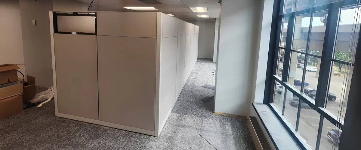 A wide shot of new office cubicle walls being installed on gray carpet, with unpacked boxes and a large window showing cars outside.