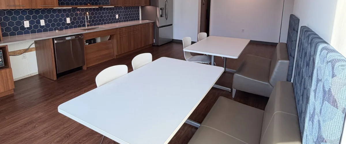 A modern common area with a kitchen, two dining tables, white chairs, and gray lounge seating. A modern common area with a kitchen, two dining tables, white chairs, and gray lounge seating.