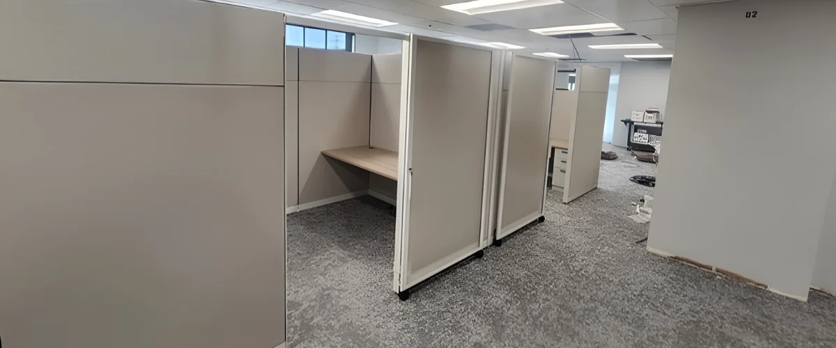 An empty office hallway with several beige privacy cubicles, one of which has a light wood desk installed. An empty office hallway with several beige privacy cubicles, one of which has a light wood desk installed.
