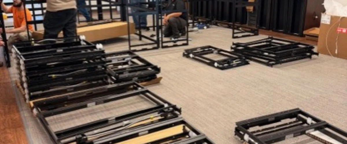 Several workers assemble black metal display frames on a light beige carpet, with cardboard boxes nearby.