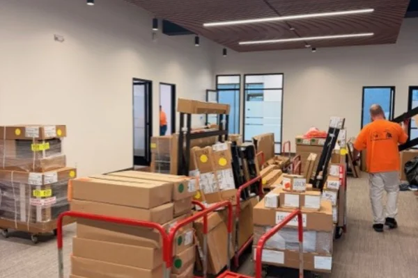 An office space filled with stacked cardboard boxes on red moving carts and a worker in an orange shirt.