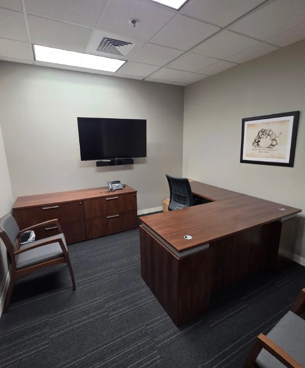 A modern office space with a large wooden L-shaped desk, a flat-screen TV mounted on the wall, and grey carpet.
