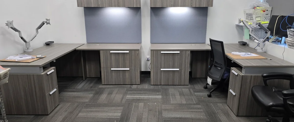 Two new, modern L-shaped office desks with light wood grain, filing cabinets, and overhead lights, ready for use. Two new, modern L-shaped office desks with light wood grain, filing cabinets, and overhead lights, ready for use.