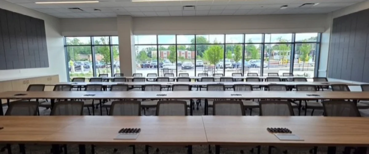 A wide shot of a modern, empty conference room with rows of tables and chairs, large windows overlook a parking lot.