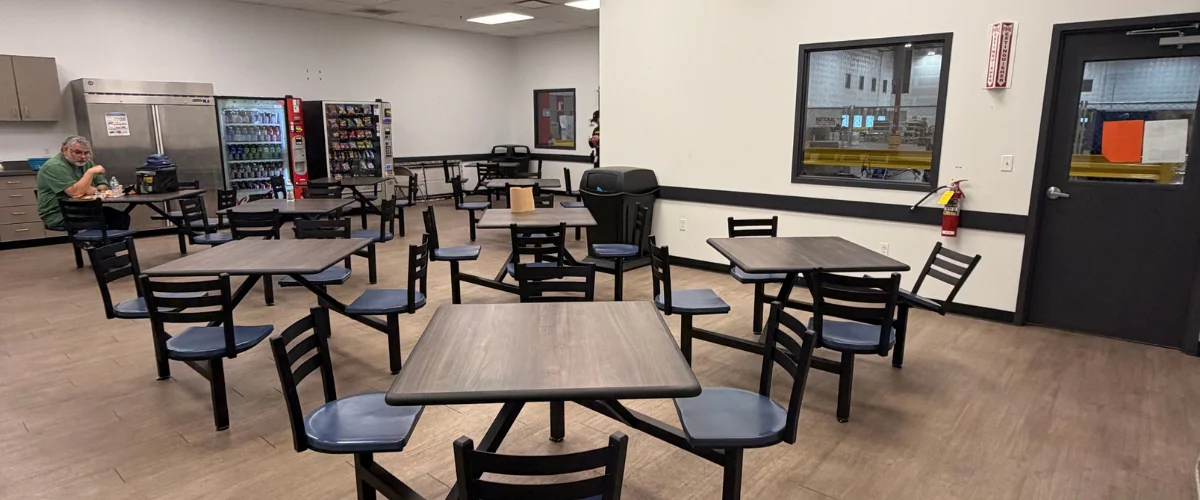 A man eats lunch in a spacious industrial breakroom with tables, vending machines, and a large refrigerator.