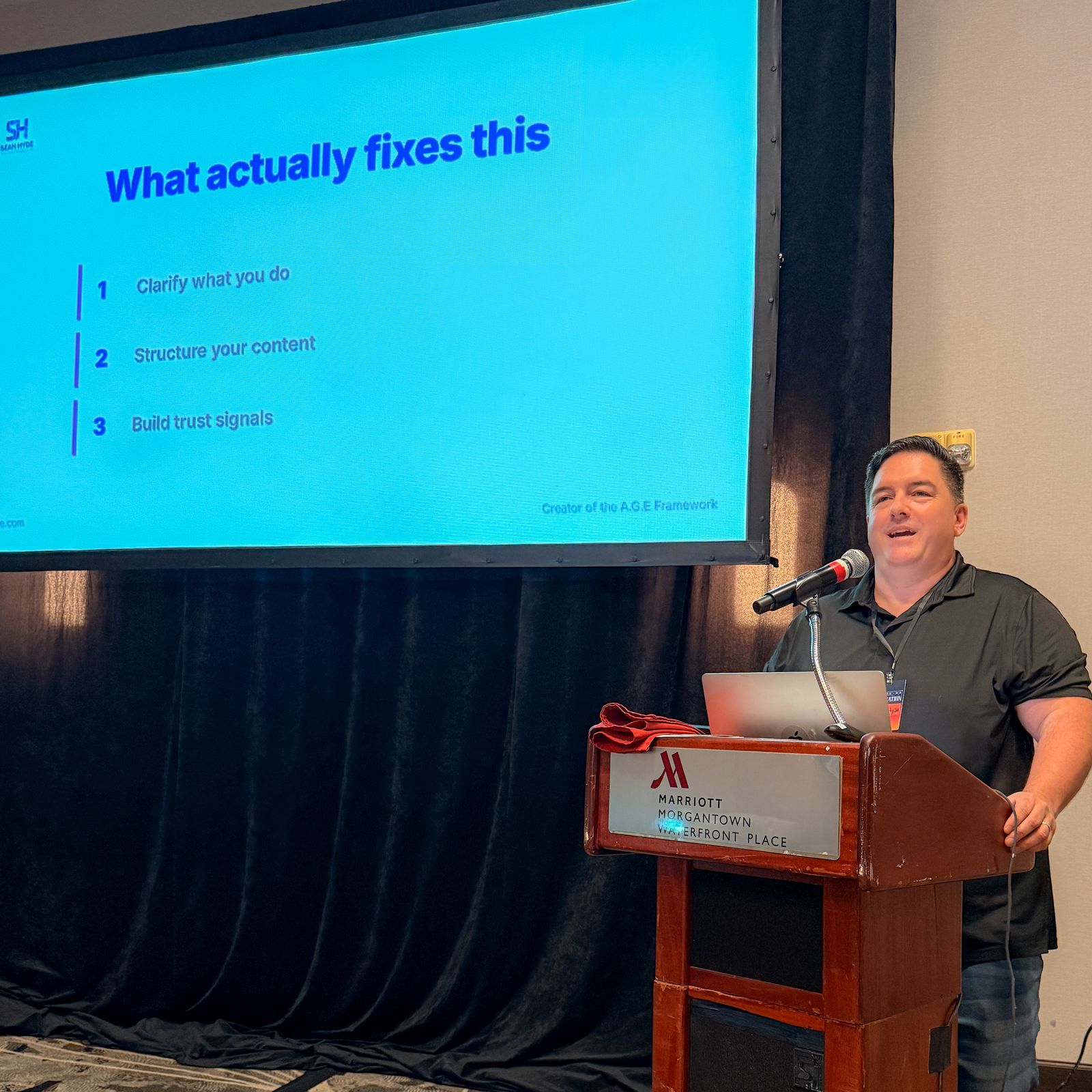 Podium at the Morgantown Marriott with Sean Hyde&rsquo;s &ldquo;What actually fixes this&rdquo; slide on screen