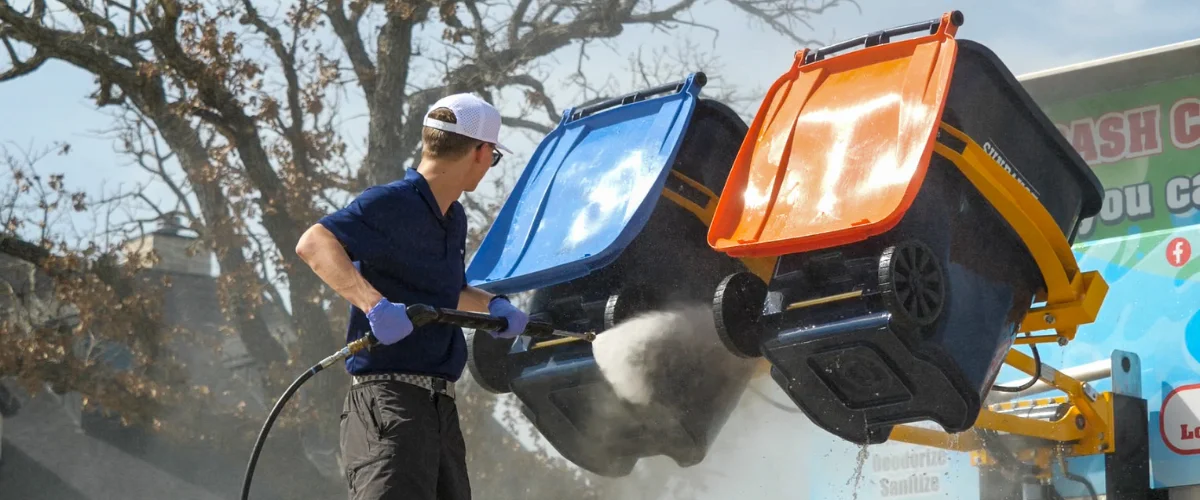 A man in uniform pressure washing a blue-lidded and orange-lidded trash can mounted on a blue truck.