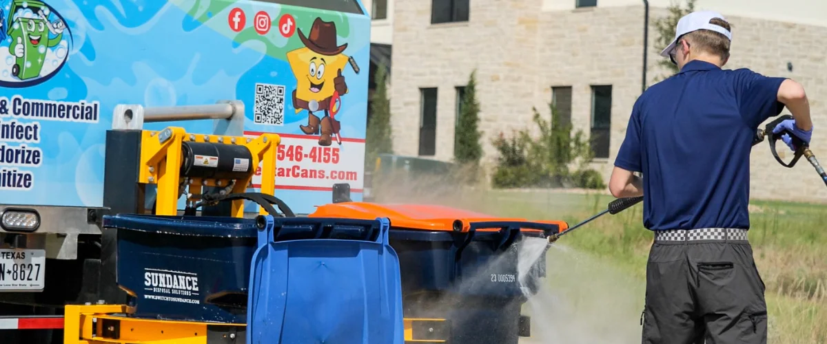 A man in a blue shirt pressure washes a dark blue and orange trash can behind a blue garbage truck, with a building in background.
