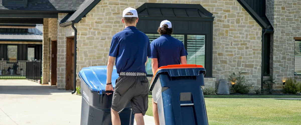 Two men in blue shirts and white caps rolling blue and black trash bins with orange lids down a driveway.