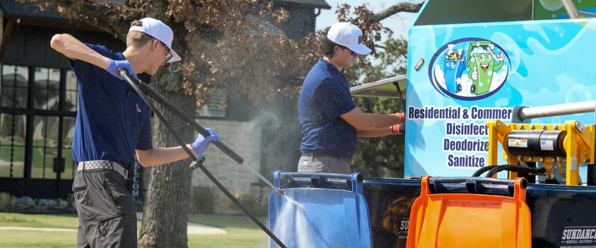 Two men power washing trash cans outdoors, a blue and an orange one, next to a service truck.