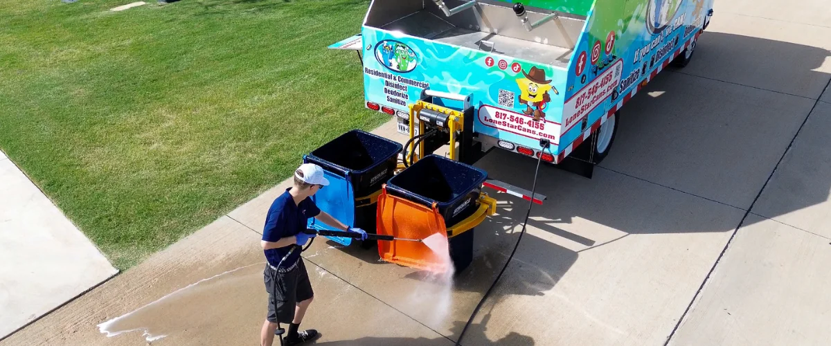 A man in blue uniform power washing an orange trash bin, next to a blue garbage cleaning truck. A man in blue uniform power washing an orange trash bin, next to a blue garbage cleaning truck.