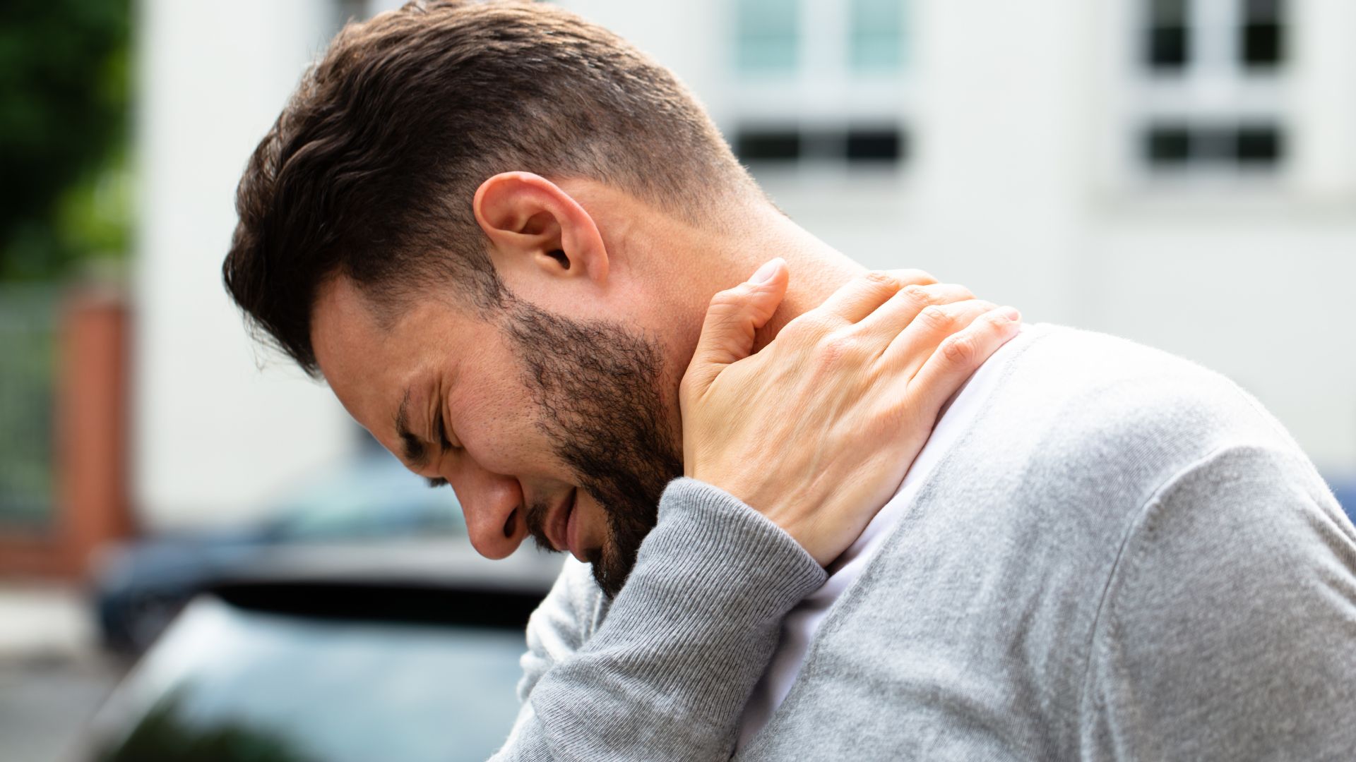 Man experiencing neck pain, holding his neck in discomfort, with a pained expression, wearing a grey sweater outdoors.